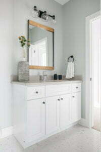 Custom made white bathroom vanity with grey counter tops, a wood mirror, a vase with flowers, and a chrome faucet.