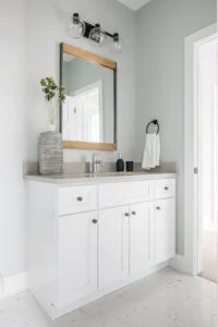 Custom made white bathroom vanity with grey counter tops, a wood mirror, a vase with flowers, and a chrome faucet.